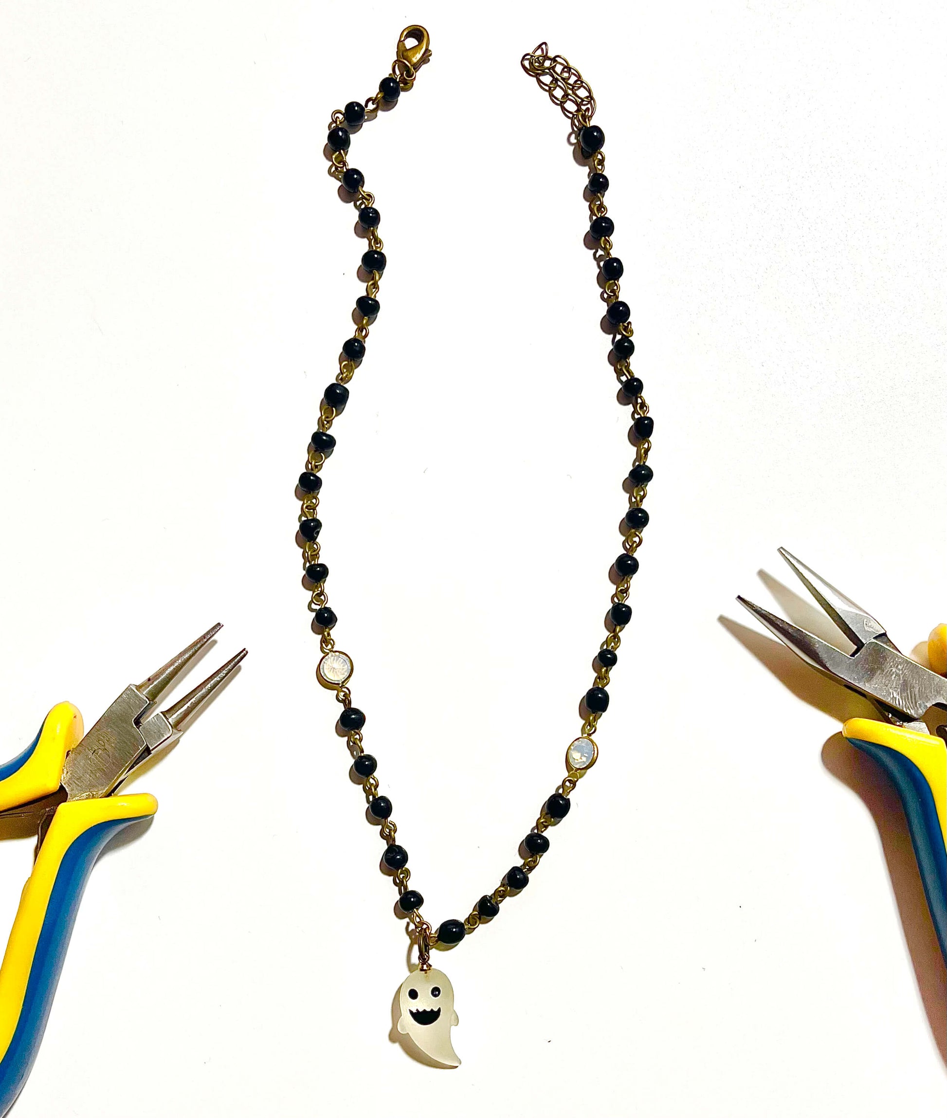 Black bead necklace with white ghost charm on white background with jewelry  making tools. 