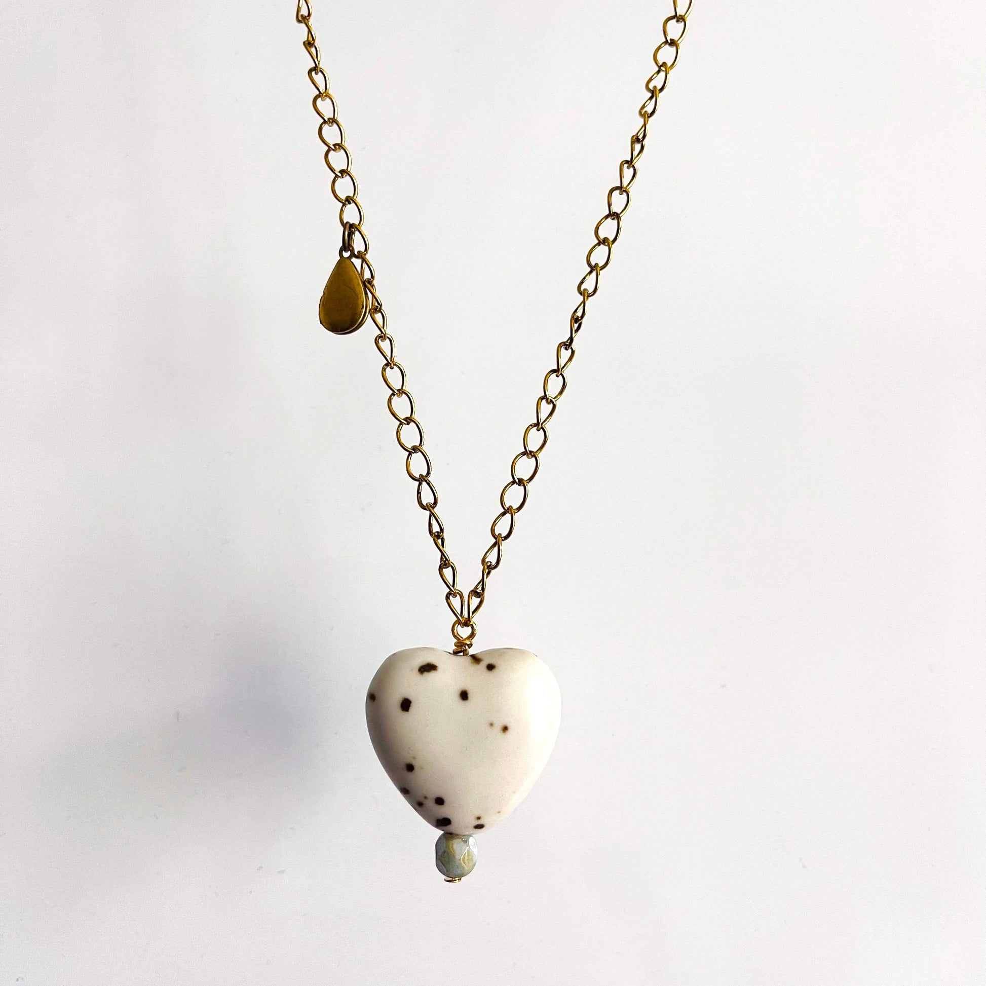 White ceramic heart pendant with brown spots and pink glass bead on a yellow brass chain with teardrop locket. 