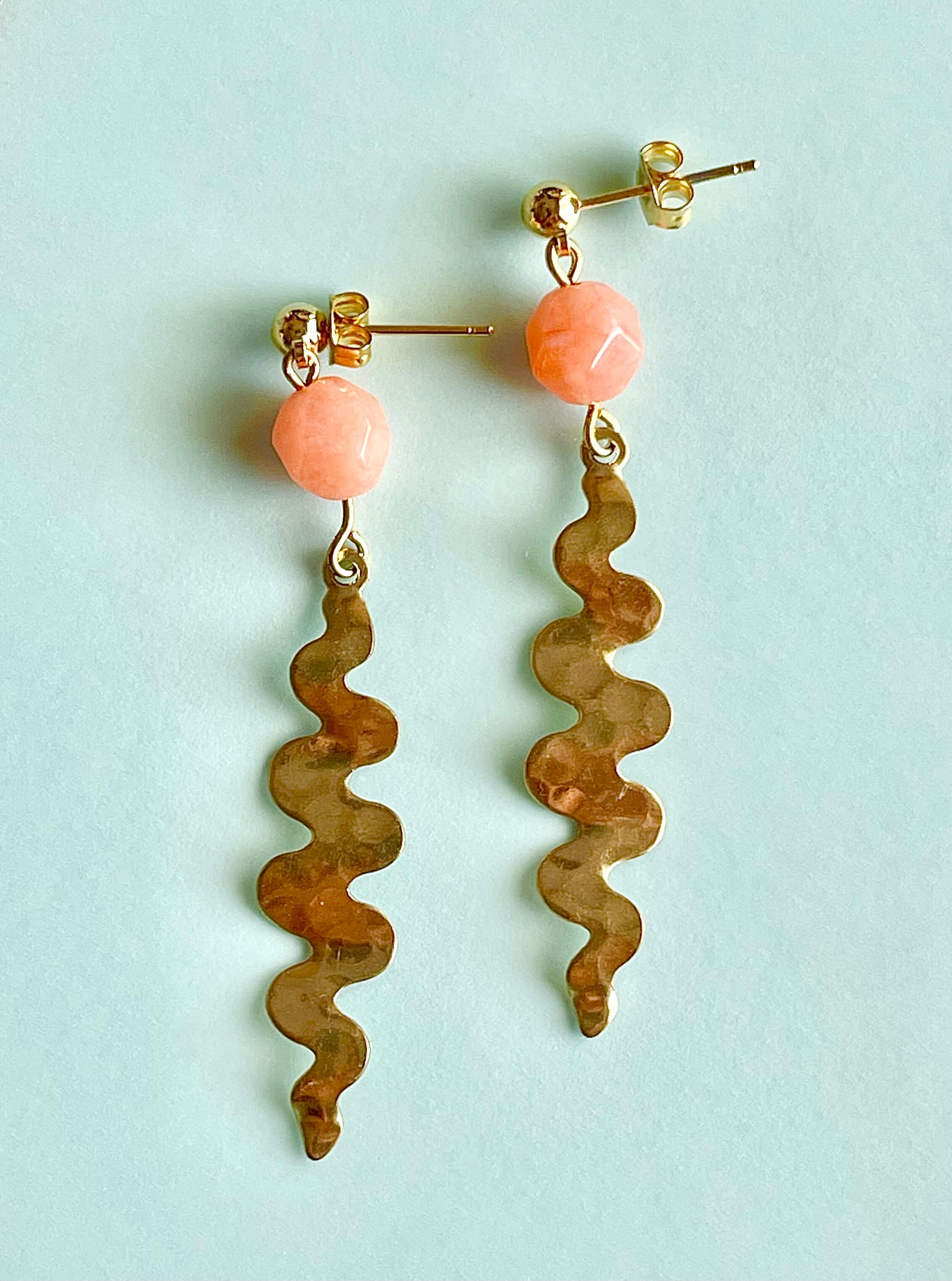 Yellow brass post earrings with wavy dangling pendants and orange faceted glass beads on a light blue background