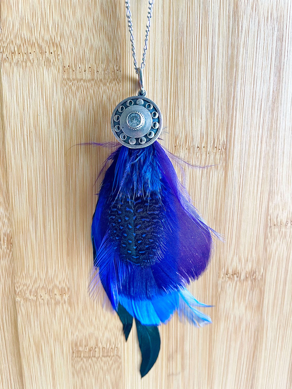 Blue and purple feathers with silver moon phase pendant on a silver chain with a wood backdrop