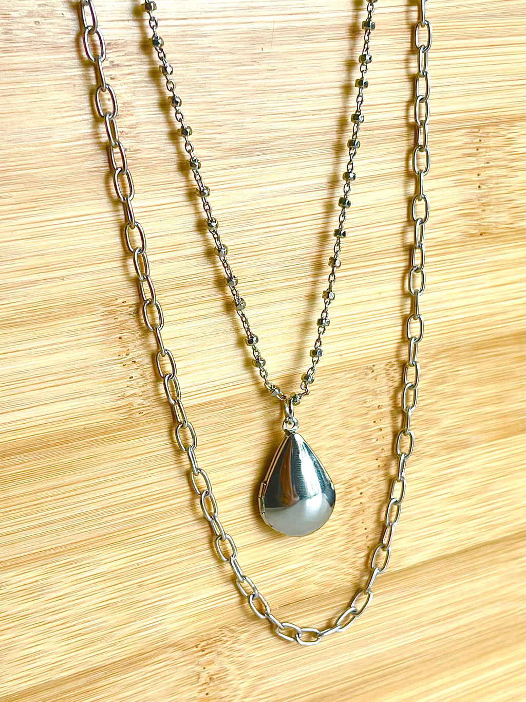 Two chains with a silver teardrop shaped locket on wood backdrop