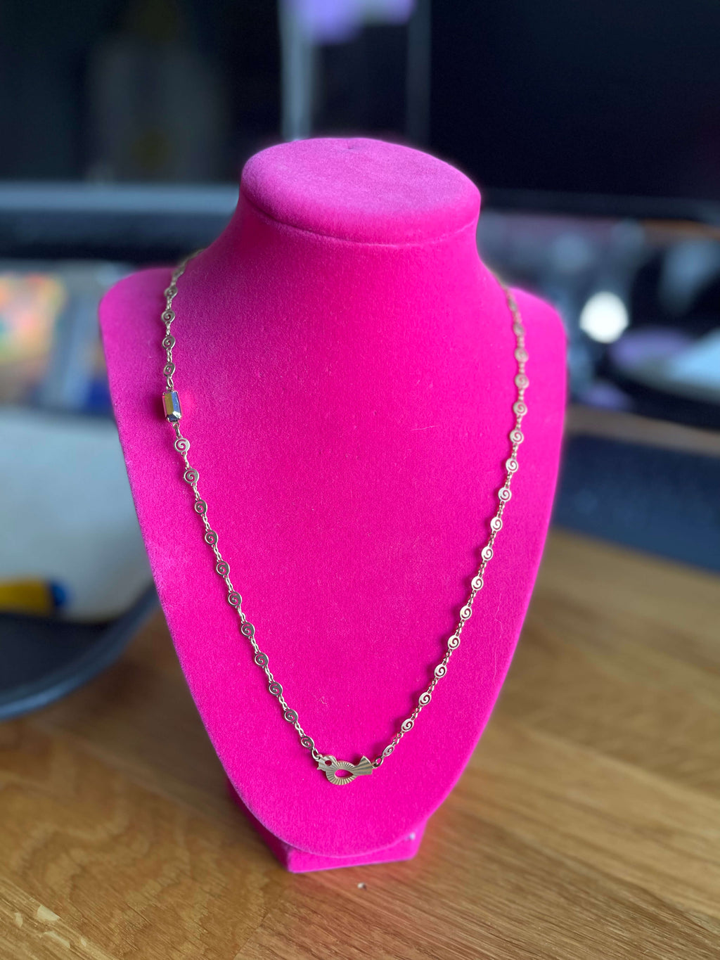 Yellow brass necklace with a swirl link chain, retro bird pendant and fuchsia crystal connector. Necklace is on a pink neck form with blurred background.  