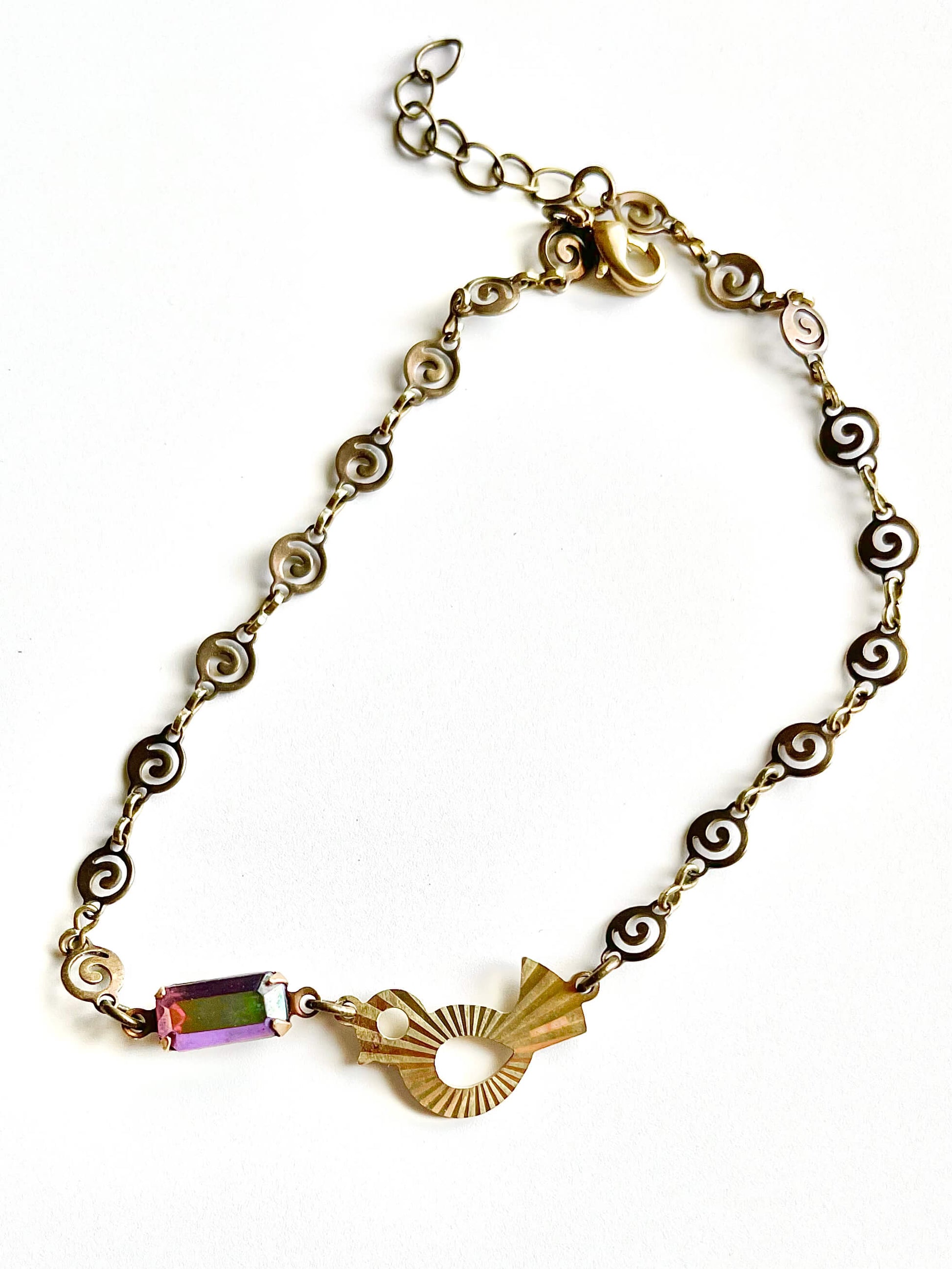Yellow brass swirl link chain with patterned bird charm and fuchsia crystal bracelet set against white backdrop. 