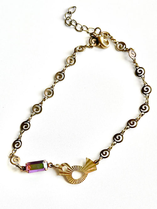 Yellow brass swirl link chain with patterned bird charm and fuchsia crystal bracelet set against white backdrop. 