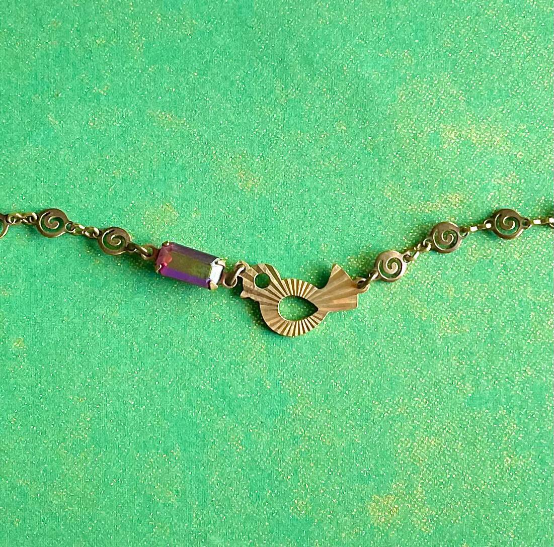 Yellow brass swirl link chain with patterned bird charm and fuchsia crystal bracelet set against green backdrop. 