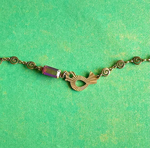 Yellow brass swirl link chain with patterned bird charm and fuchsia crystal bracelet set against green backdrop. 