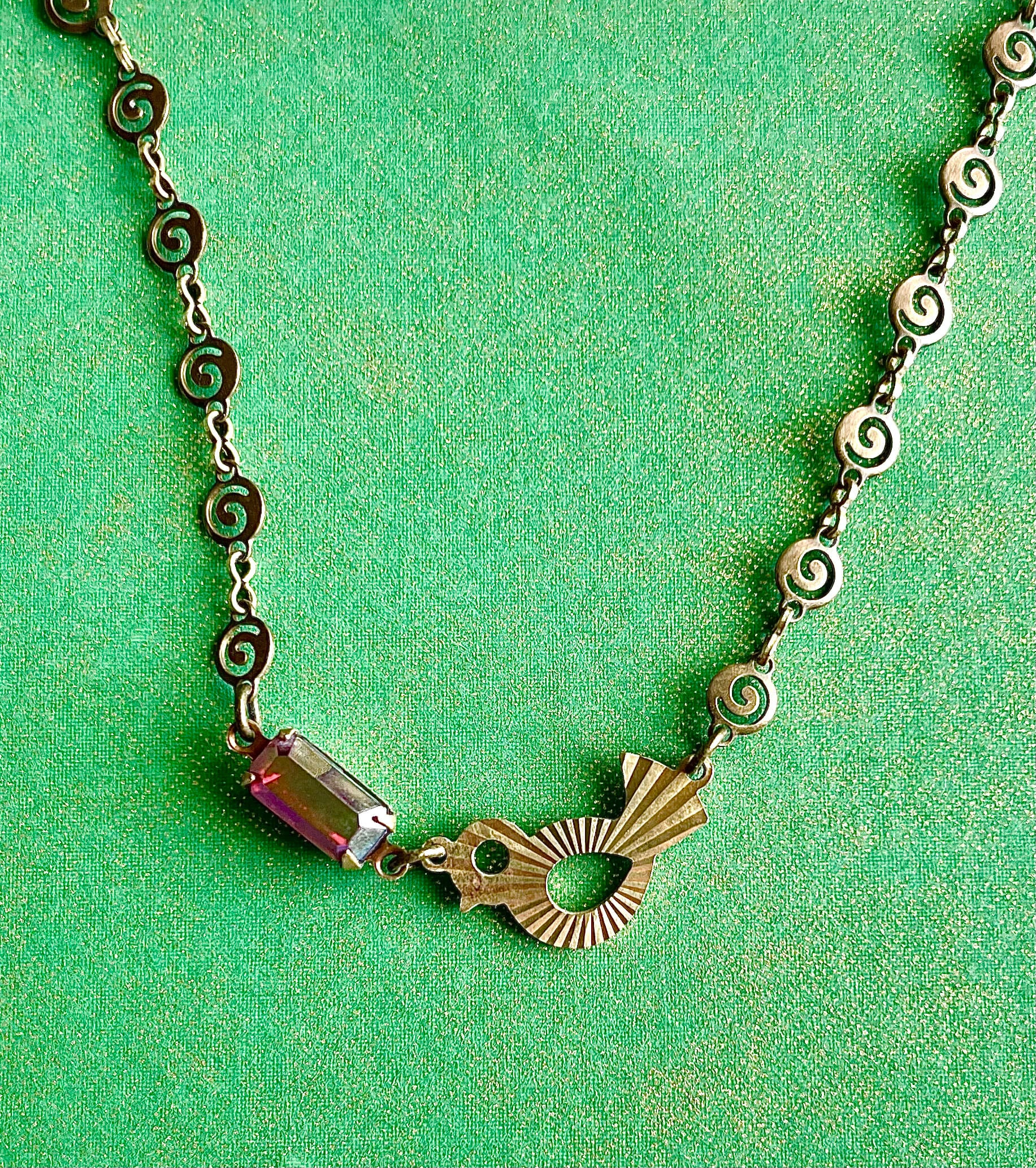 Yellow brass swirl link chain with patterned bird charm and fuchsia crystal bracelet set against green backdrop. 