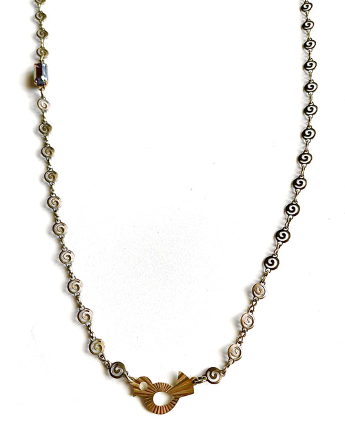 Yellow brass swirl link chain with patterned bird charm and fuchsia crystal necklace set against green backdrop. 