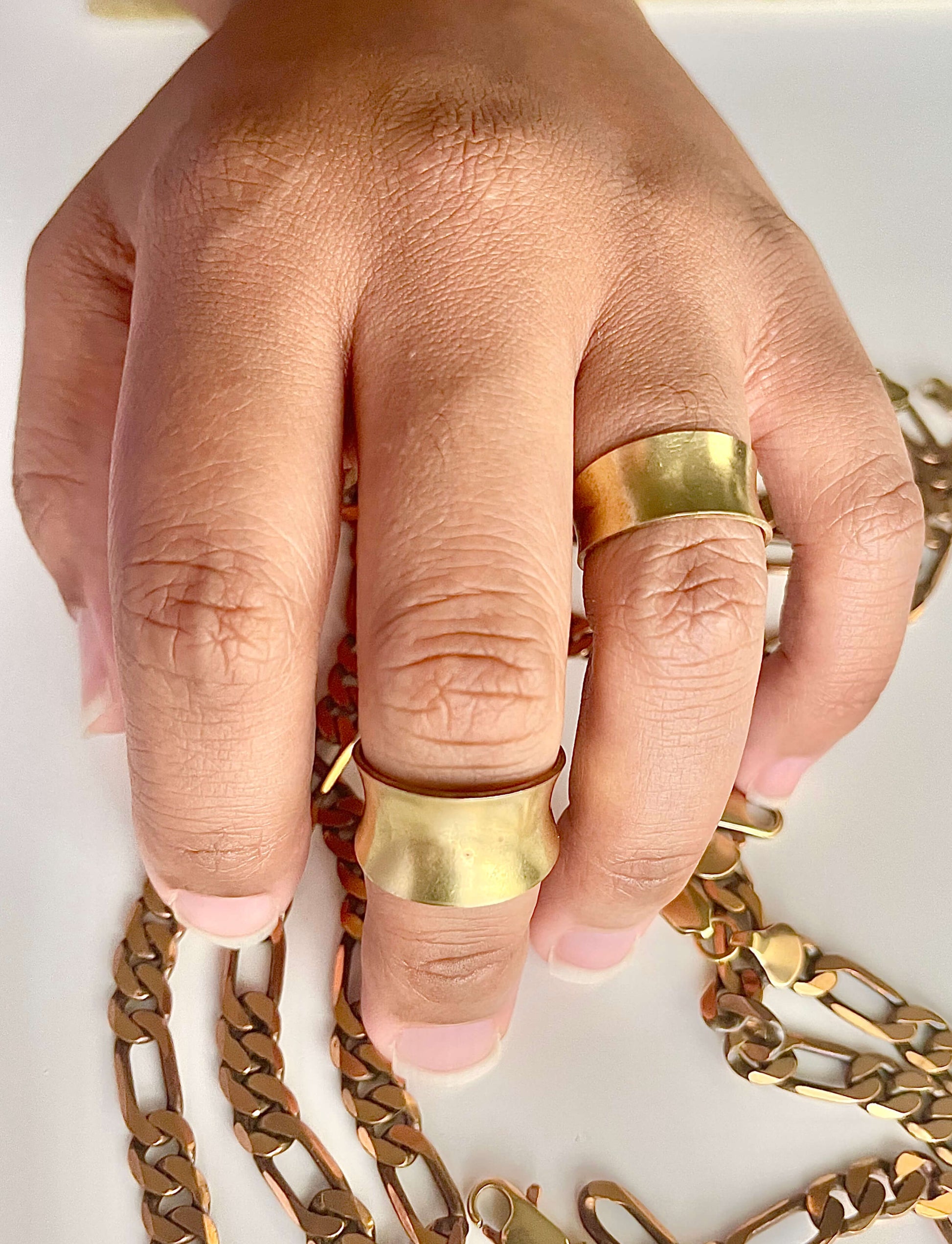 A hand wearing a wide yellow brass ring on one finger, and a narrow yellow brass ring on another finger. 